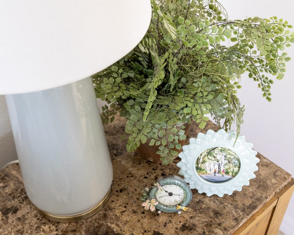 A decorated nightstand in the Primary Bedroom with a Sea Foam Blue Lamp, coordinating picture frames and a plant bring beauty to this functional space.